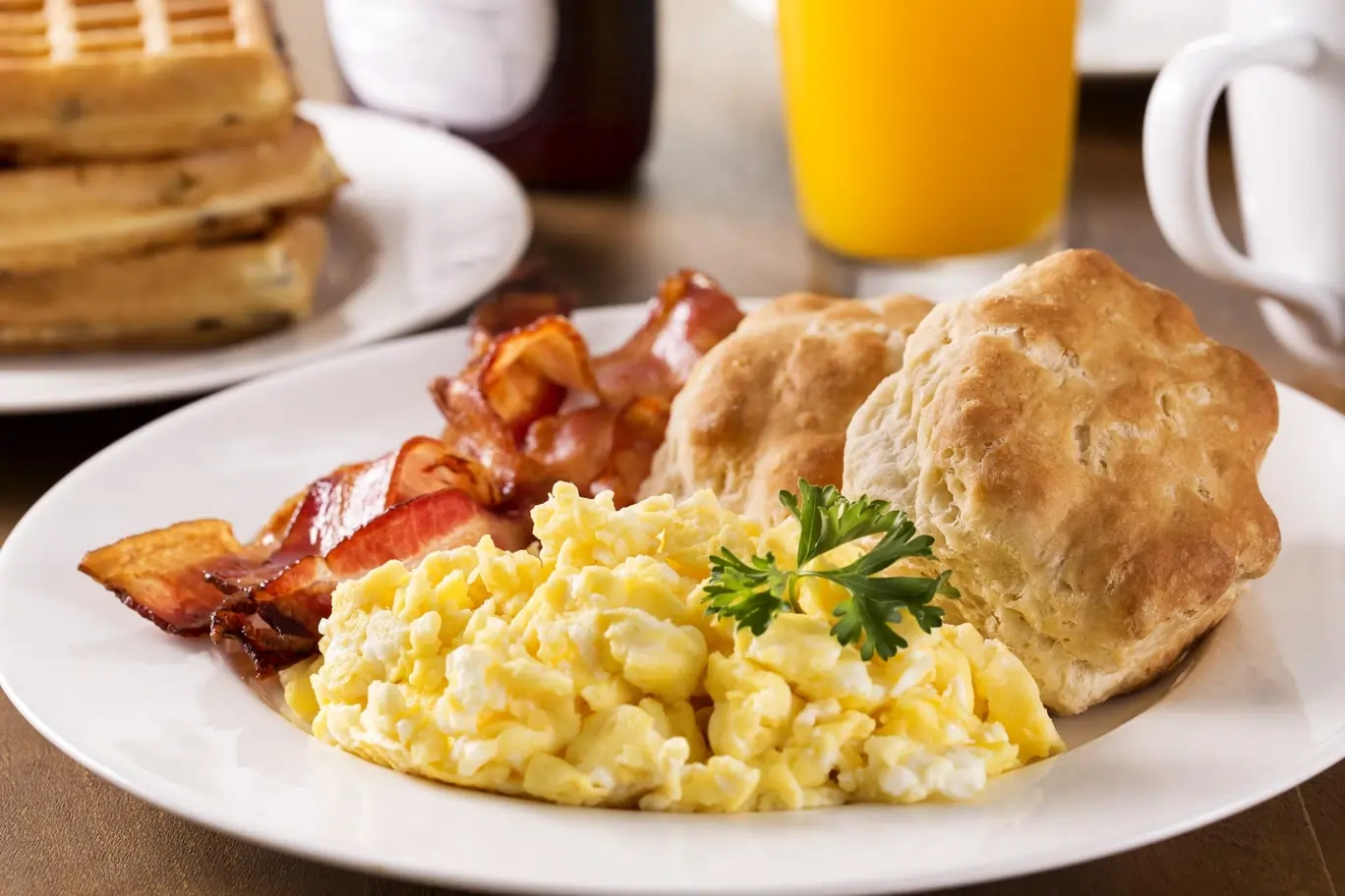 Scrambled eggs, bacon, and biscuits served with orange juice and waffles in the background